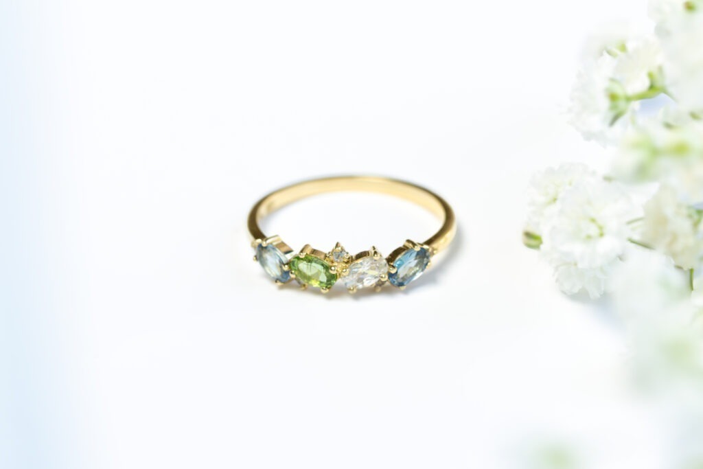 White background with flowers on the right hand side with a gold band morse code ring with 4 stones in blue, green, white